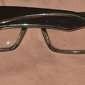 Pu&ograve; includere: Occhiali Ray-Ban neri con montatura rettangolare e lenti trasparenti. Il logo Ray-Ban &egrave; visibile sulle aste. Gli occhiali sono appoggiati su uno sfondo rosa.