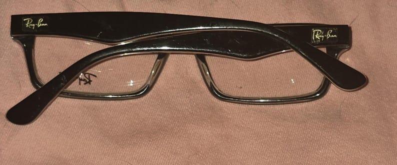 Pu&ograve; includere: Occhiali Ray-Ban neri con montatura rettangolare e lenti trasparenti. Il logo Ray-Ban &egrave; visibile sulle aste. Gli occhiali sono appoggiati su uno sfondo rosa.