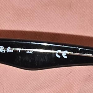 Pu&ograve; includere: Primo piano dell'asta di occhiali da sole neri. Il logo Ray-Ban &egrave; visibile in bianco, insieme al testo "MAD" e "CE". L'asta &egrave; nera con una cerniera argentata.