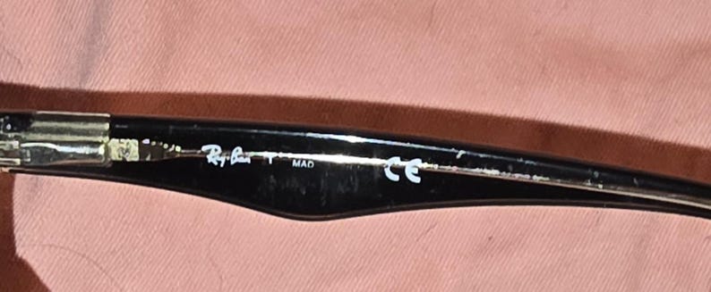 Pu&ograve; includere: Primo piano dell'asta di occhiali da sole neri. Il logo Ray-Ban &egrave; visibile in bianco, insieme al testo "MAD" e "CE". L'asta &egrave; nera con una cerniera argentata.
