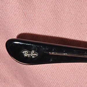Pu&ograve; includere: Primo piano dell'asta di una montatura di occhiali da sole Ray-Ban neri. Il logo Ray-Ban &egrave; visibile in lettere argentate sull'asta. La montatura &egrave; posta su uno sfondo di tessuto rosa.