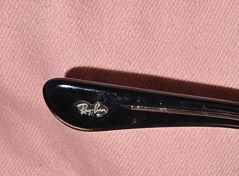 Pu&ograve; includere: Primo piano dell'asta di una montatura di occhiali da sole Ray-Ban neri. Il logo Ray-Ban &egrave; visibile in lettere argentate sull'asta. La montatura &egrave; posta su uno sfondo di tessuto rosa.