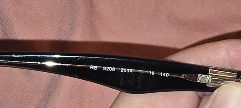 Pu&ograve; includere: Primo piano dell'asta di occhiali da sole neri. Il testo "RB 5208 2034 18 140" &egrave; stampato in bianco all'interno dell'asta. L'asta &egrave; tenuta da una persona.