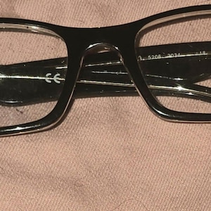 Pu&ograve; includere: Occhiali Ray-Ban neri con montatura rettangolare. Gli occhiali hanno lenti trasparenti e il logo Ray-Ban &egrave; visibile sulla lente destra. Le aste degli occhiali sono anch'esse nere e presentano il logo Ray-Ban.