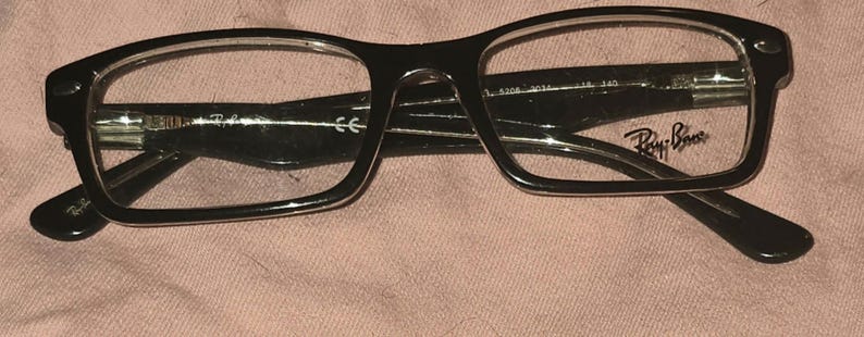 Pu&ograve; includere: Occhiali Ray-Ban neri con montatura rettangolare. Gli occhiali hanno lenti trasparenti e il logo Ray-Ban &egrave; visibile sulla lente destra. Le aste degli occhiali sono anch'esse nere e presentano il logo Ray-Ban.