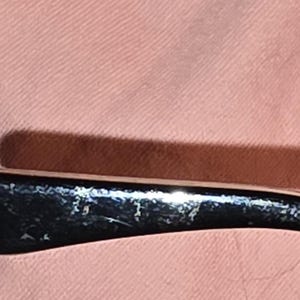 Pu&ograve; includere: Primo piano di un'asta di occhiali nera con un accento argentato. L'asta ha un design curvo e mostra segni di usura. Lo sfondo &egrave; di colore rosa chiaro.