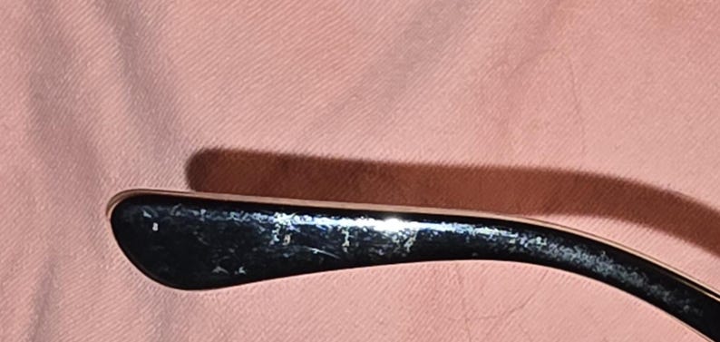 Pu&ograve; includere: Primo piano di un'asta di occhiali nera con un accento argentato. L'asta ha un design curvo e mostra segni di usura. Lo sfondo &egrave; di colore rosa chiaro.