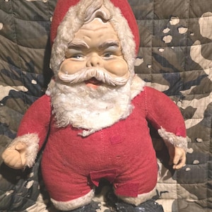 May include: Vintage Santa Claus doll with a red suit, hat, and black boots. The doll has a white beard and a slightly grumpy expression. The doll is made of fabric and has a painted face.