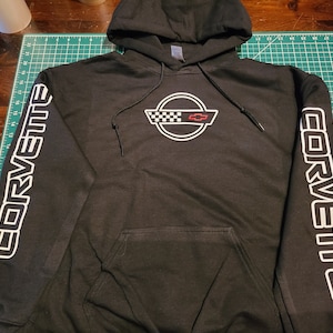 May include: Black hooded sweatshirt with white "Corvette" text on the sleeves and a white Corvette logo on the front. The logo features a checkered flag and a red Chevrolet emblem.
