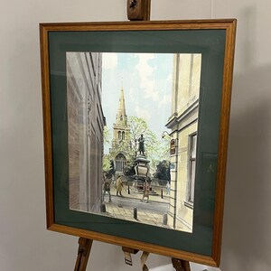 May include: A framed watercolor painting depicting a town square with a central statue and a church spire in the background. The artwork is housed in a dark green mat and a wooden frame, displayed on a wooden tripod easel. The scene includes figures.