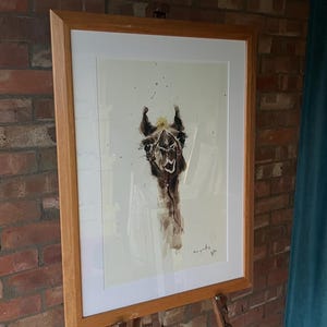 May include: A framed watercolor painting of a llama with a light brown body and a white background. The llama has a light brown nose and a dark brown eye. The painting is signed "Sue Gardner" in the bottom right corner.