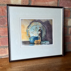 May include: Framed watercolour painting of a still life with a blue and gold plate, a blue ceramic pot, a bottle of wine, and two oranges on a table.