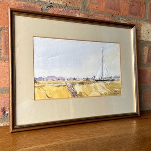 May include: Framed watercolor painting of a sailboat on a waterway. The artwork features a white sailboat with a tall mast, a wooden pier, and a yellow field. The painting is set against a light blue sky and is framed in a brown wooden frame.