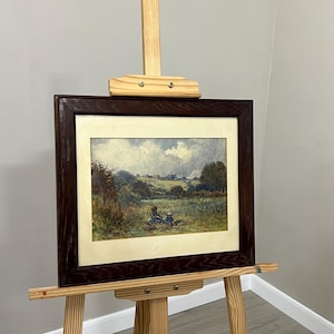 May include: A framed landscape painting with a dark brown wooden frame is displayed on a wooden easel. The painting depicts a field with figures, trees, and a cloudy sky. The easel is made of light-colored wood.