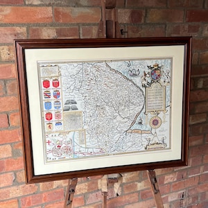 Puede incluir: Un mapa antiguo enmarcado del condado de Kent, Inglaterra, con una ilustración detallada de la geografía y las ciudades de la región. El mapa se muestra en un caballete de madera.