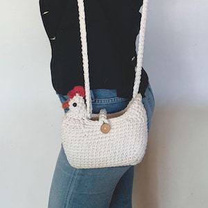 Puede incluir: Un bolso de hombro de ganchillo blanco con forma de pollo, con una cresta roja y un cierre de bot&oacute;n de madera.