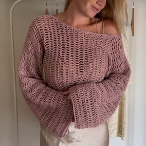 Op de afbeelding: Een oudroze gehaakte trui met een wijde boothals, lange uitlopende mouwen en een verticaal opengewerkt patroon. Het kledingstuk wordt gedragen over een lichtbeige satijnen rok met een gladde, glanzende afwerking en een eenvoudige zoom.