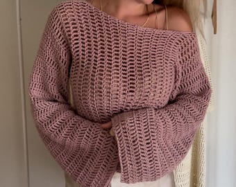 May include: A dusty rose crochet sweater featuring a wide boat neckline, long flared sleeves, and an open-knit vertical stitch pattern. The garment is worn over a light beige satin skirt with a smooth, lustrous finish and a simple hemline.