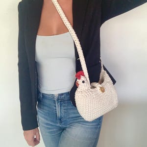Puede incluir: Un bolso de mano blanco de ganchillo con forma de pollo, con una cresta roja y un bot&oacute;n marr&oacute;n. El bolso se lleva al hombro con una correa blanca.
