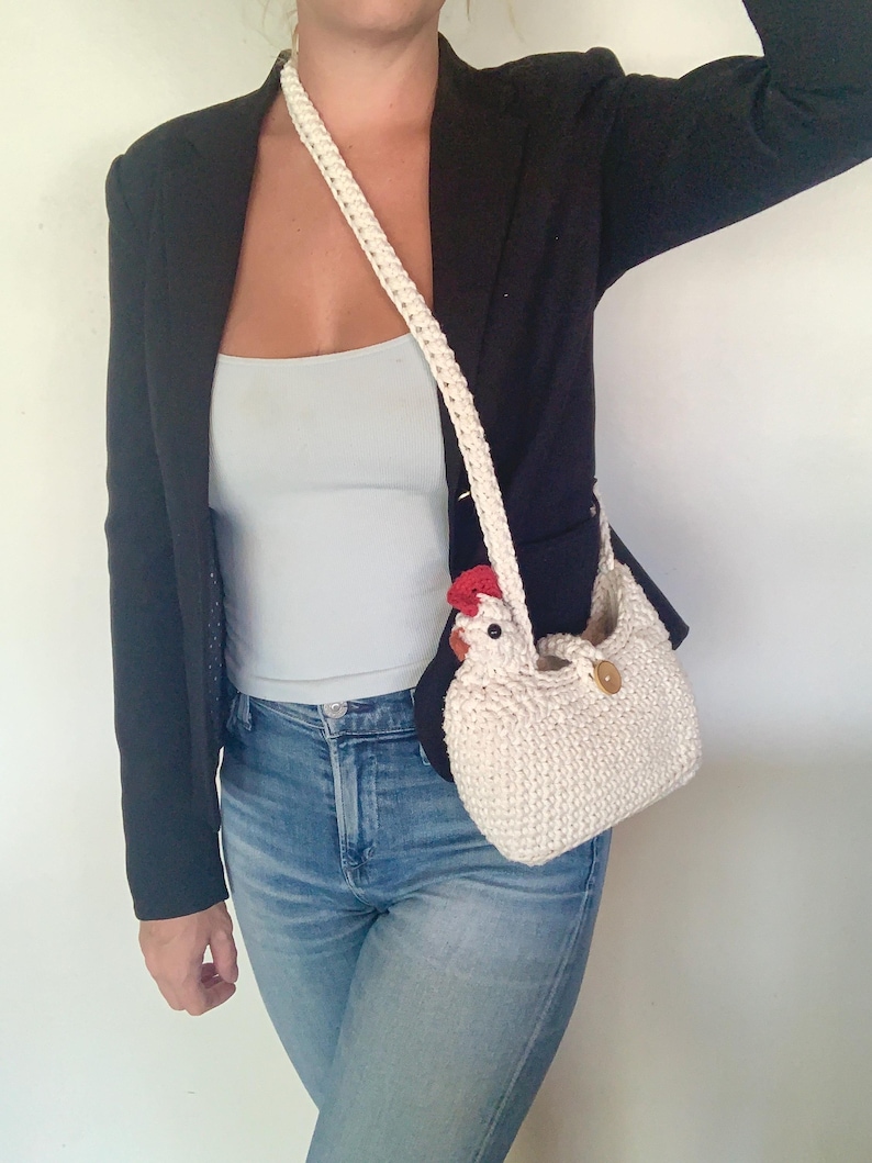 Puede incluir: Un bolso de mano blanco de ganchillo con forma de pollo, con una cresta roja y un bot&oacute;n marr&oacute;n. El bolso se lleva al hombro con una correa blanca.