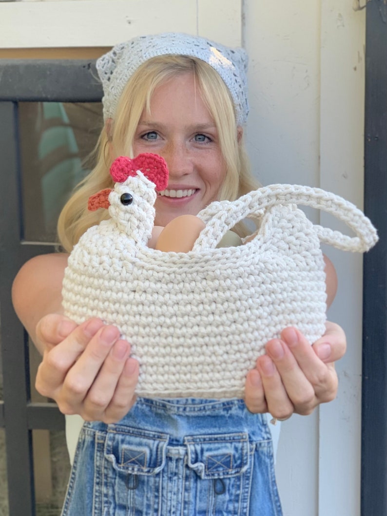 Puede incluir: Una cesta de crochet con forma de pollo con una cresta roja y un asa de crochet blanco. La cesta est&aacute; llena de huevos y la est&aacute; sujetando una persona que lleva un mono vaquero azul y una diadema de crochet azul claro.