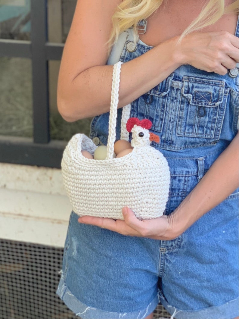 Puede incluir: Una cesta de crochet blanca con forma de pollo con una cresta roja y un pico marr&oacute;n. La cesta est&aacute; llena de huevos y la est&aacute; sujetando una persona que lleva un mono vaquero.