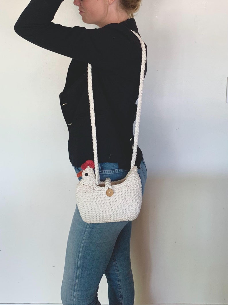 Puede incluir: Un bolso de hombro de ganchillo blanco con forma de pollo, con una cresta roja y un cierre de bot&oacute;n de madera.