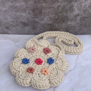 May include: A crocheted cream-colored flower-shaped bag with a long strap. The bag has colorful embroidered flowers in shades of pink, blue, and orange.