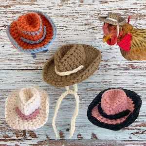 May include: Four crocheted cowboy hats in different colors: orange, brown, white, and pink. The hats are all handmade and have a classic cowboy hat shape.