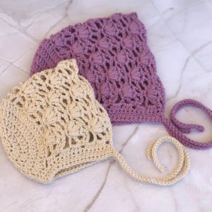Könnte beinhalten: Zwei gehäkelte Babyhauben, eine in hellbeige und die andere in staubigem Rosa. Beide Hauben haben ein zartes, spitzenartiges Muster und sind mit passenden Bändern gebunden.