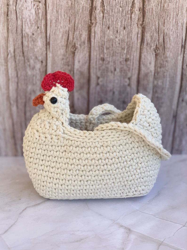 Puede incluir: Una cesta de crochet con forma de pollo con una cresta roja y ojos negros. La cesta est&aacute; hecha de hilo blanco y tiene un asa.