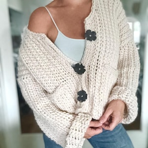 May include: A cream-colored knitted cardigan with black flower-shaped buttons. The cardigan has a relaxed fit and long sleeves.