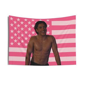 May include: A pink American flag tapestry featuring a smiling person. The flag design includes white stars on a pink background and pink stripes. The person is shirtless, wearing black shorts, and has dark hair.