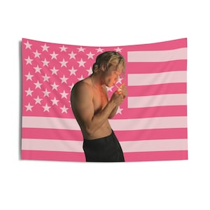 May include: Pink American flag tapestry with a person lighting a cigarette. The flag features white stars on a pink field and pink and white stripes. The person is wearing black pants.