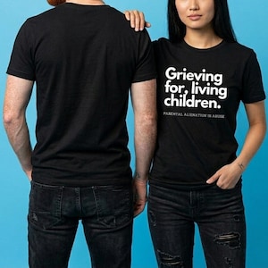 Op de afbeelding: Twee mensen in zwarte t-shirts en donkere jeans staan tegen een blauwe achtergrond. Het t-shirt aan de rechterkant heeft de witte tekst "Grieving for, living children." en "PARENTAL ALIENATION IS ABUSE".