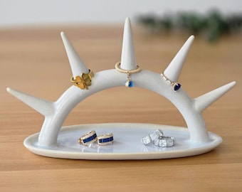 May include: A white ceramic ring holder in the shape of antlers, with a small oval base. Several rings with gold and blue accents are displayed on the holder, including a four-leaf clover ring and a ring with a blue gemstone.