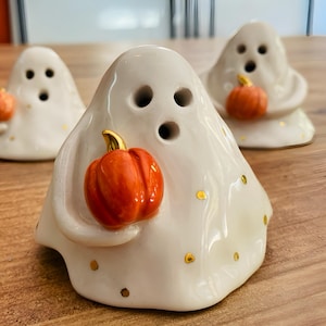 May include: Three white ceramic ghost figurines with gold accents. Each ghost is holding a small orange pumpkin.
