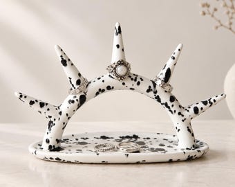 May include: A white ceramic ring holder with a black paint splatter pattern. The holder features an arched base with five upward-pointing cones for displaying rings. Three rings are placed on the cones, and two rings rest on the oval base.
