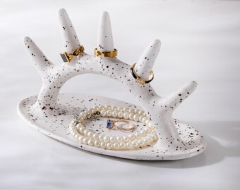 May include: White ceramic ring holder shaped like a branch with black speckles. The holder has four prongs for rings and a small dish for jewelry. A pearl necklace and two rings are displayed on the holder.
