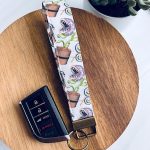 May include: White fabric wristlet key fob with a repeating pattern of a purple, green, and brown plant design. The wristlet has a bronze-colored metal clasp and a key ring. A black car key fob is next to the wristlet.