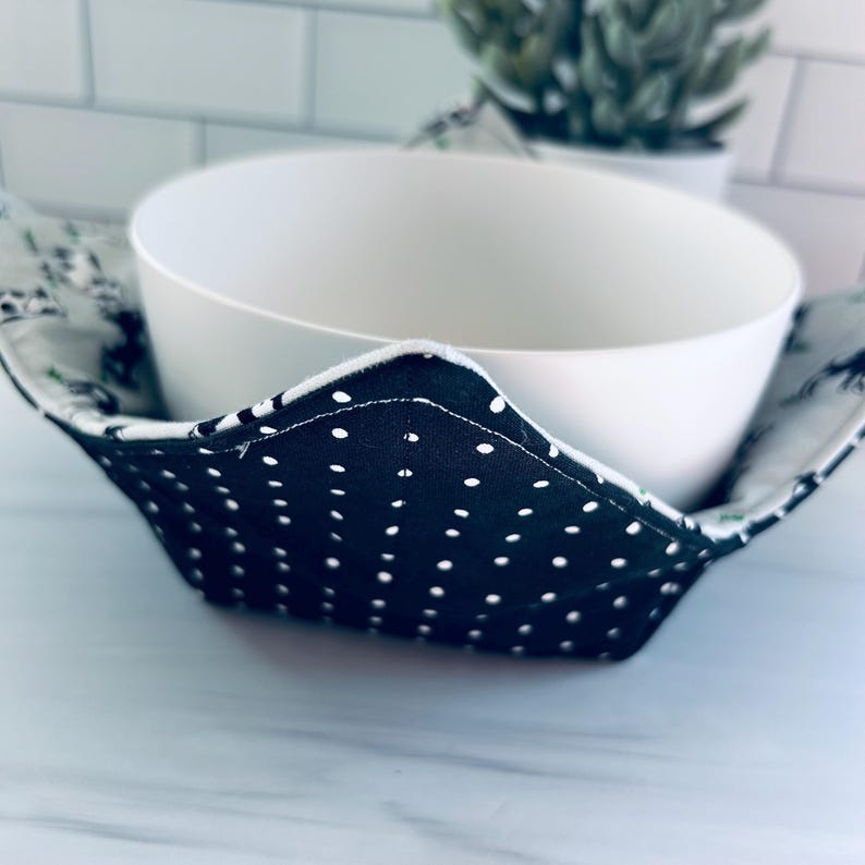 May include: A white ceramic bowl sits inside a black and white polka dot fabric bowl cozy. The cozy has a white trim and a floral pattern on the inside. The background includes a white tile wall and a small potted plant.