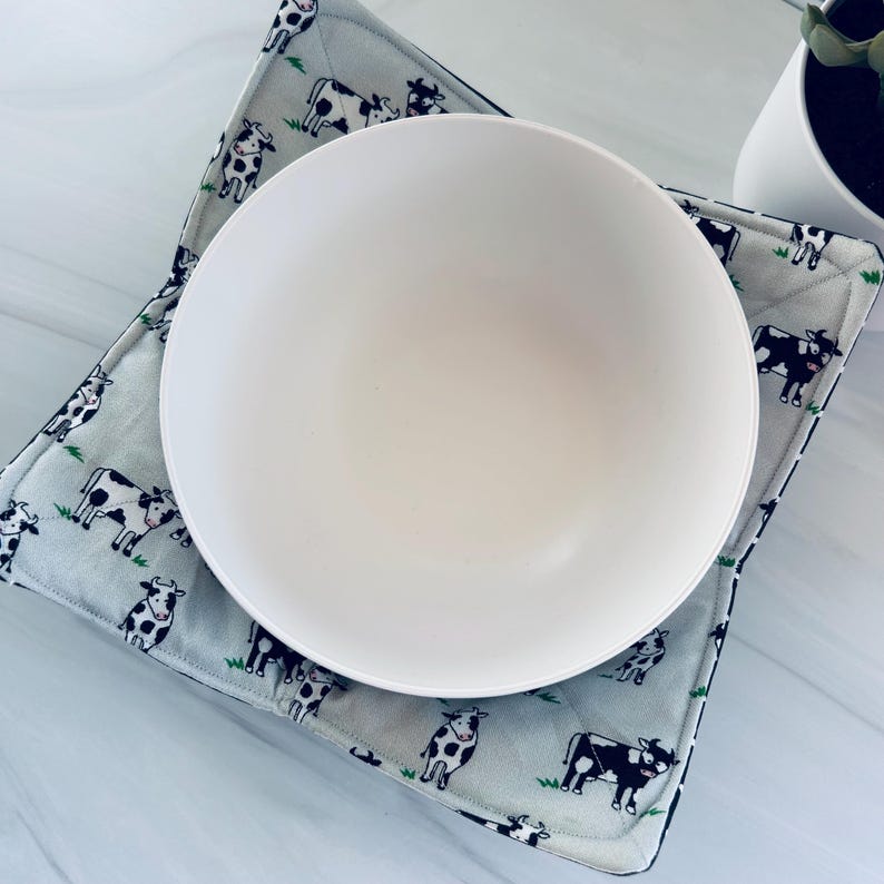 May include: A white ceramic bowl sits atop a square, quilted pot holder. The pot holder features a light blue background with a repeating pattern of black and white cows. The edges of the pot holder are black.