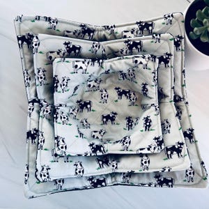 May include: A set of three quilted bowl cozies in varying sizes. The cozies feature a light gray background with a repeating pattern of black and white cows and green grass. The edges are trimmed with a dark gray binding.