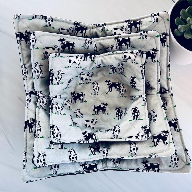 May include: A set of three quilted bowl cozies in varying sizes. The cozies feature a light gray background with a repeating pattern of black and white cows and green grass. The edges are trimmed with a dark gray binding.