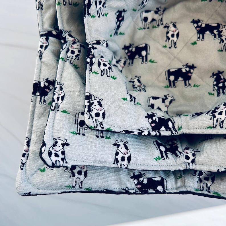 May include: A light gray quilted blanket featuring a repeating pattern of black and white cows grazing on green grass. The blanket is folded, showcasing the detailed stitching and the cow print design. The edges are neatly finished.