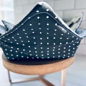 May include: A dark gray bowl cozy with white polka dots. The bowl cozy is made of fabric and has a square shape with folded corners. The cozy is sitting on a small, round, wooden stool with metal legs.