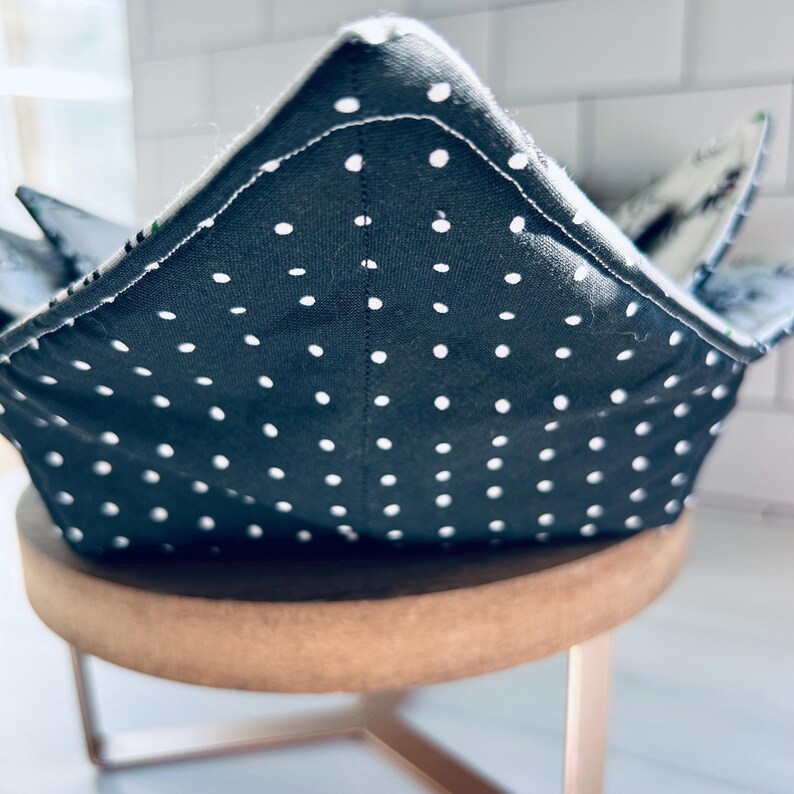 May include: A dark gray bowl cozy with white polka dots. The bowl cozy is made of fabric and has a square shape with folded corners. The cozy is sitting on a small, round, wooden stool with metal legs.