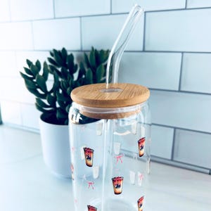 Puede incluir: Un vaso de vidrio transparente con tapa de bambú y pajita de vidrio curvada. El vaso está decorado con un patrón de ilustraciones de bebidas rojas y blancas y lazos rosas. Una pequeña planta en maceta está en el fondo.