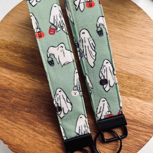 May include: Two key fobs with a light green background and a repeating pattern of white ghosts holding trick-or-treat buckets and jack-o'-lanterns. Each fob has a black metal ring and hardware.