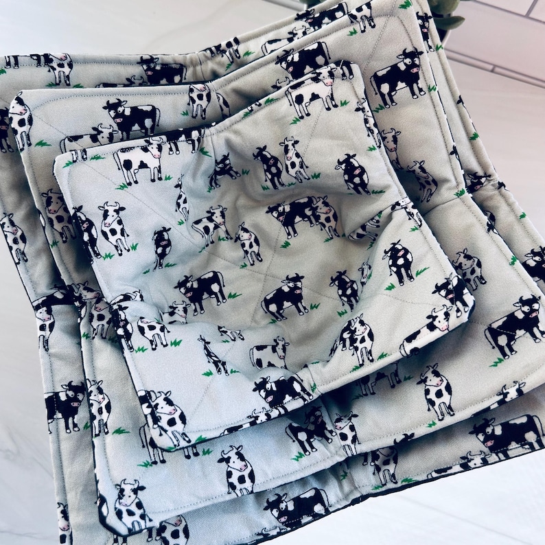 May include: A stack of four gray bowl cozies with a repeating pattern of black and white cows on a green field. The cozies are quilted and have a black border.